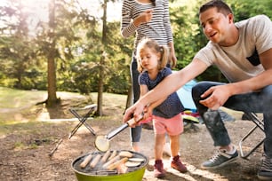 Beautiful family enjoying camping holiday in forest. Father cooking meat on barbecue grill.