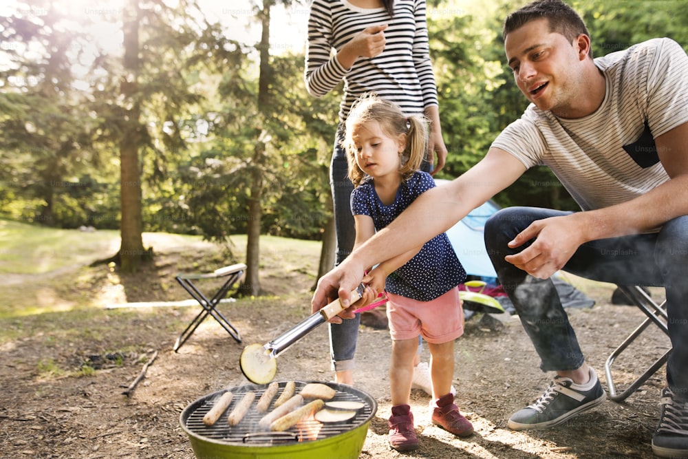 Beautiful family enjoying camping holiday in forest. Father cooking meat on barbecue grill.