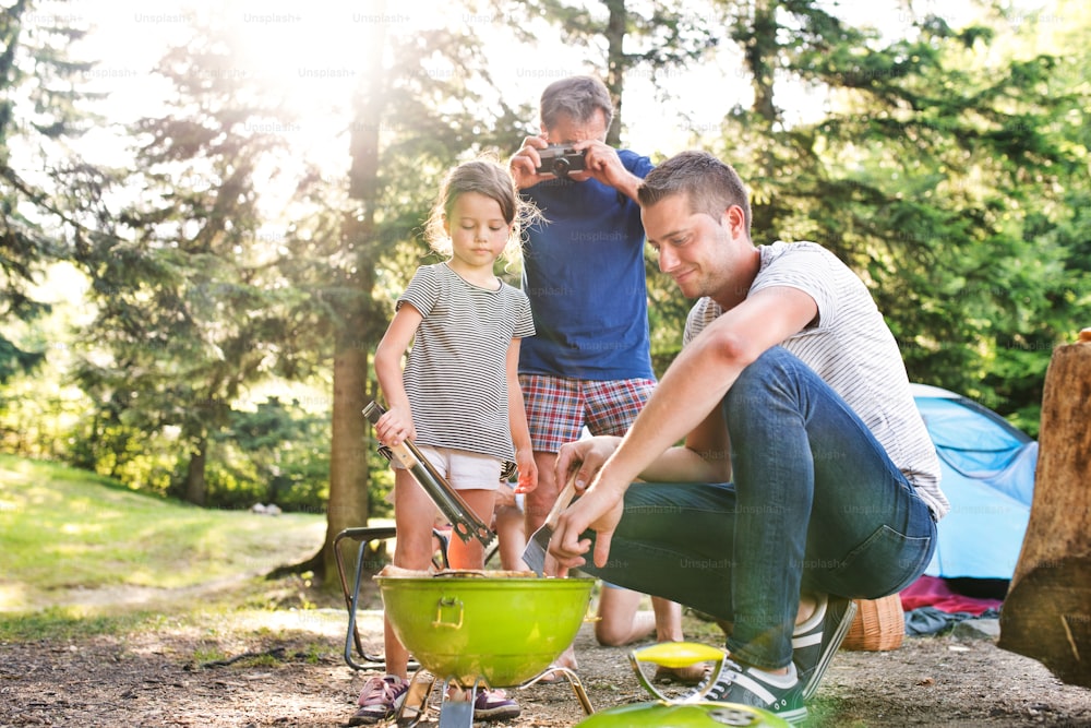 Beautiful family enjoying camping holiday in forest. Father cooking meat on barbecue grill, grandfather taking picture with camera.