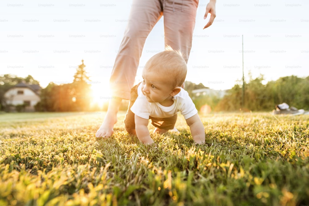 Cute baby boy with his unrecognizable mother crawling outside on the green grass.