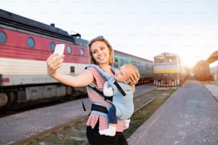 아기와 함께 기차로 여행하는 젊은 엄마. 스마트폰을 들고 있는 여자와 그녀의 아들이 기차역에 있다.