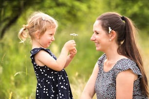 Beautiful young mother in green sunny summer nature with her cute little daughter, girl giving her daisy flower.