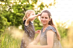 Beautiful young mother in green sunny summer nature with her cute little daughter, girl giving her daisy flower.