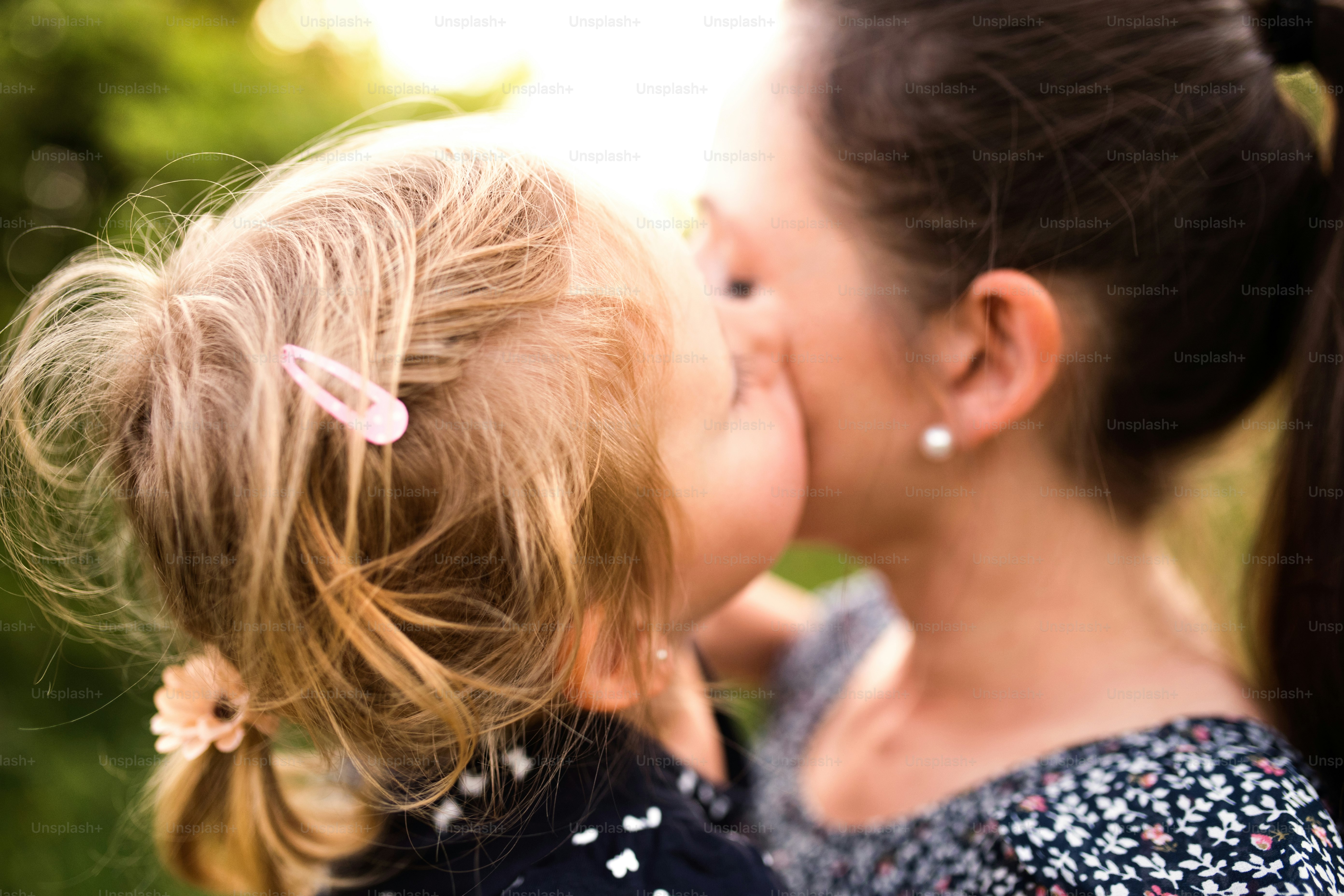 Beautiful young mother in green sunny summer nature holding her cute little daughter in the arms, girl kissing her on cheek.