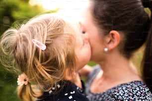 Hermosa madre joven en la naturaleza verde y soleada del verano sosteniendo a su linda hijita en los brazos, niña besándola en la mejilla.