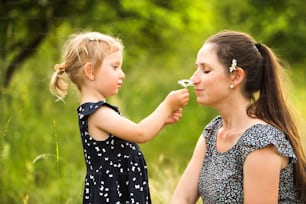 Beautiful young mother in green sunny summer nature with her cute little daughter, girl giving her daisy flower, mother smelling it.