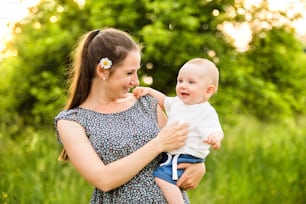 Beautiful young mother in green sunny summer nature holding baby son in the arms.