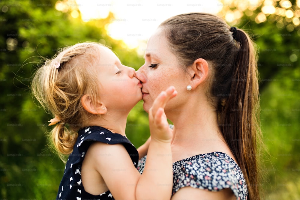 녹색 햇살이 내리쬐는 여름 자연에 있는 아름다운 젊은 어머니는 귀여운 딸을 팔에 안고 코에 키스하는 소녀.