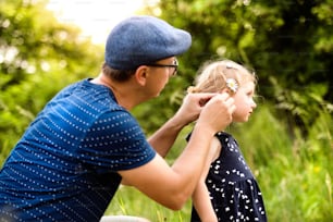 Young father with his cute little daughter spending time together outside in green summer nature doing her hairstyle.