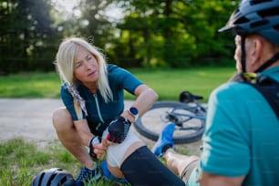 Une femme âgée aide un homme après qu’il soit tombé d’un vélo sur le sol et se soit blessé au genou, dans un parc en été.