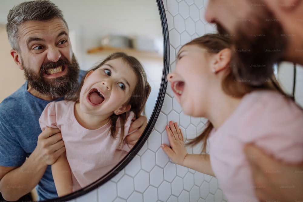 Ein Vater, der sich mit seiner kleinen Tochter amüsiert und im Badezimmer zu Hause im Spiegel Grimassen schneidet.