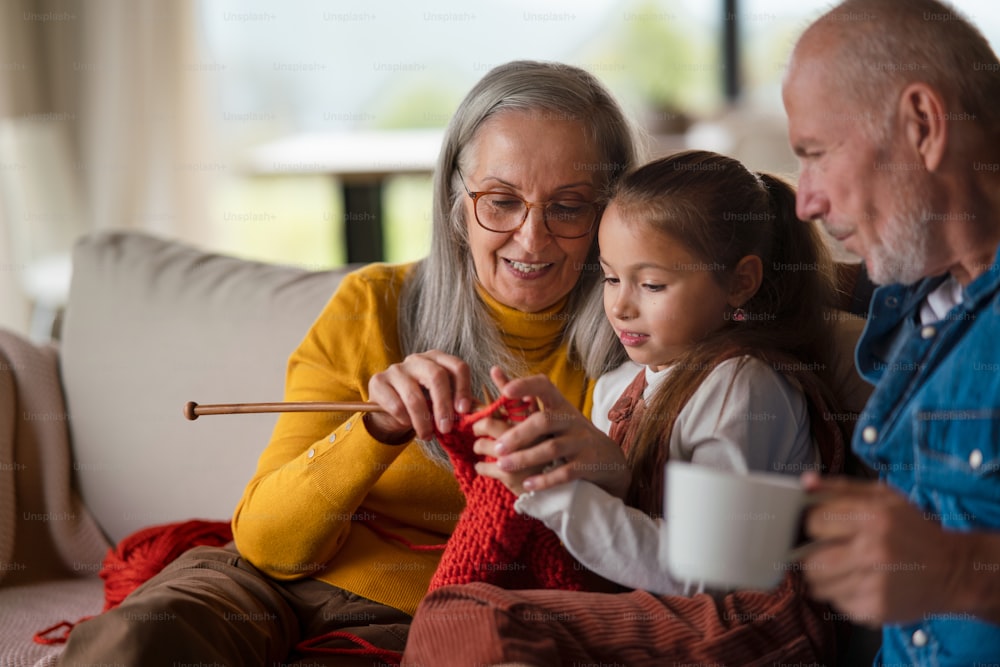 Une petite fille assise sur un canapé avec ses grands-parents et apprenant à tricoter à l’intérieur à la maison.