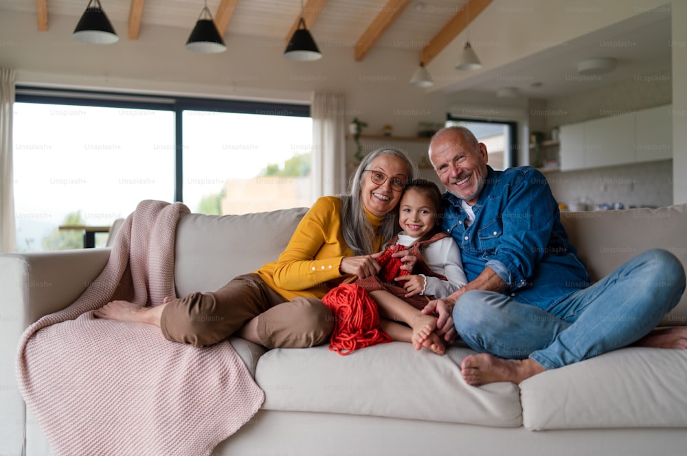 A little girl sitting on sofa with her grandparents and learning to knit indoors at home.