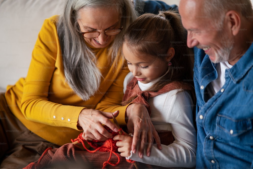 A little girl sitting on sofa with her grandparents and learning to knit indoors at home.
