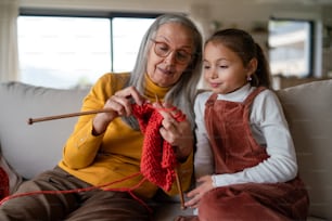 A grandmother sitting on sofa and teaching her granddaughter how to knit indoors at home.