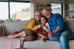 Une petite fille assise sur un canapé avec ses grands-parents et apprenant à tricoter à l’intérieur à la maison, regardant la caméra. Une grand-mère assise sur un canapé et apprenant à sa petite-fille à tricoter à l’intérieur à la maison.