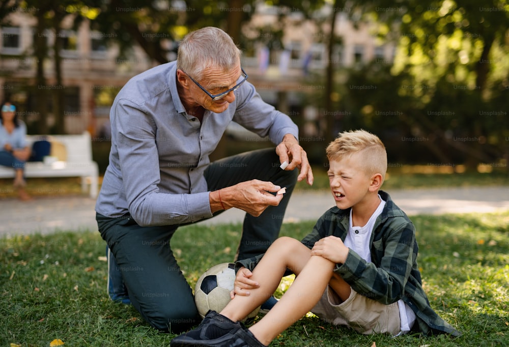 Um garotinho com a perna ferida chorando, seu avô está lhe dando gesso ao ar livre no parque.