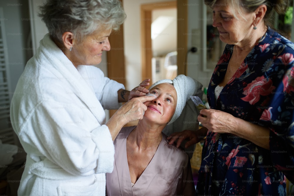 Happy senior women friends in bathrobes applying anti wrinkle patches under eyes indoors at home, selfcare concept.