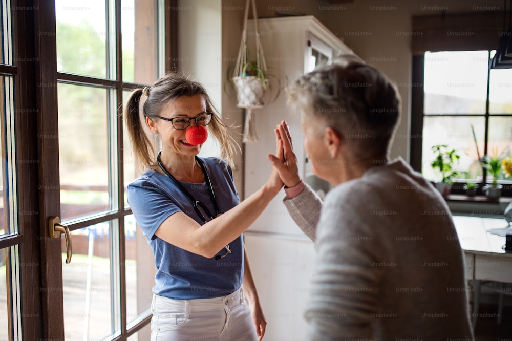 A healthcare worker or caregiver with red nose visiting senior woman indoors at home, having fun.