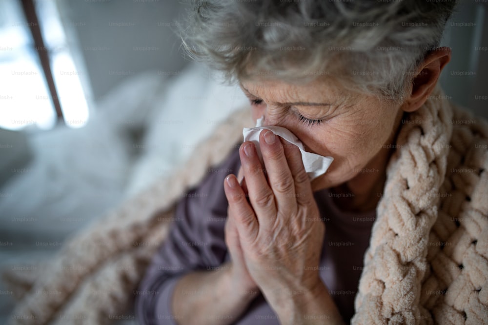 A sick senior woman in bed at home, sneezing and blowing nose.