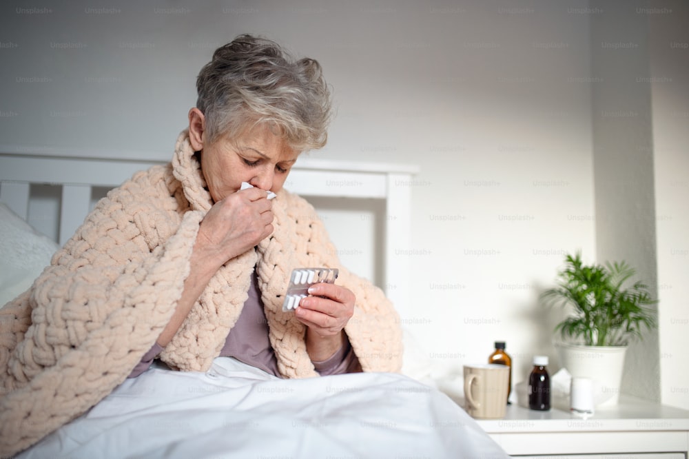 Eine kranke ältere Frau liegt zu Hause im Bett, niest und nimmt Medikamente ein.