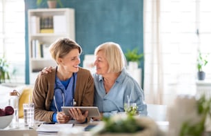 Senior woman with caregiver or healthcare worker indoors, using tablet and talking.