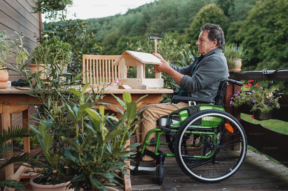 Uomo anziano in sedia a rotelle che costruisce casetta per uccelli all'aperto sulla terrazza, concetto di progetto fai-da-te.