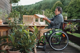 Senior man in wheelchair constructing birdhouse outdoors on terrace, diy project concept.