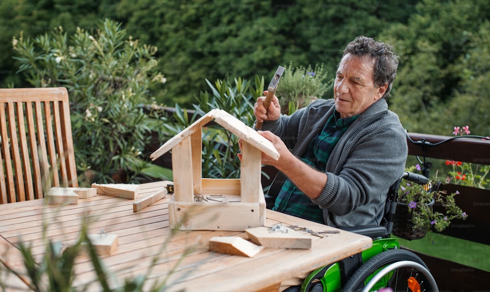 Senior man in wheelchair constructing birdhouse outdoors on terrace, diy project concept.