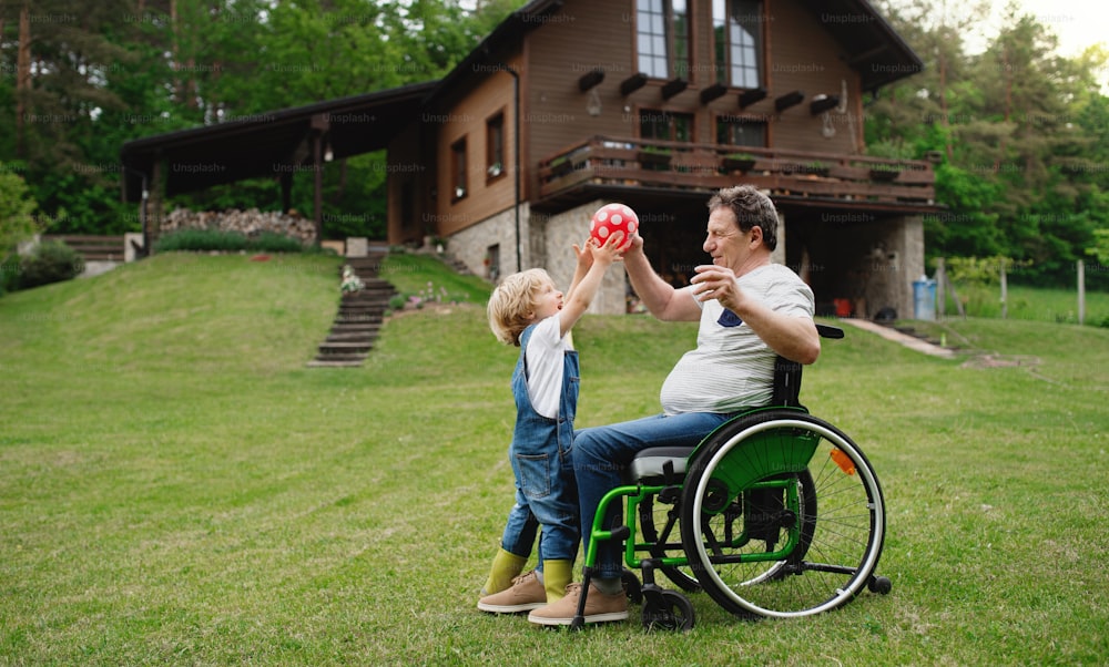 Happy small boy with senior grandfather in wheelchair in garden, playing with a ball.