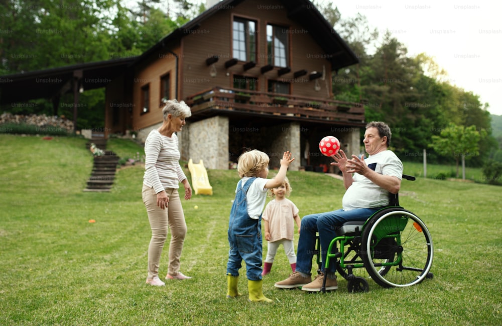 Happy small children with senior grandparents in wheelchair playing with a ball in garden.