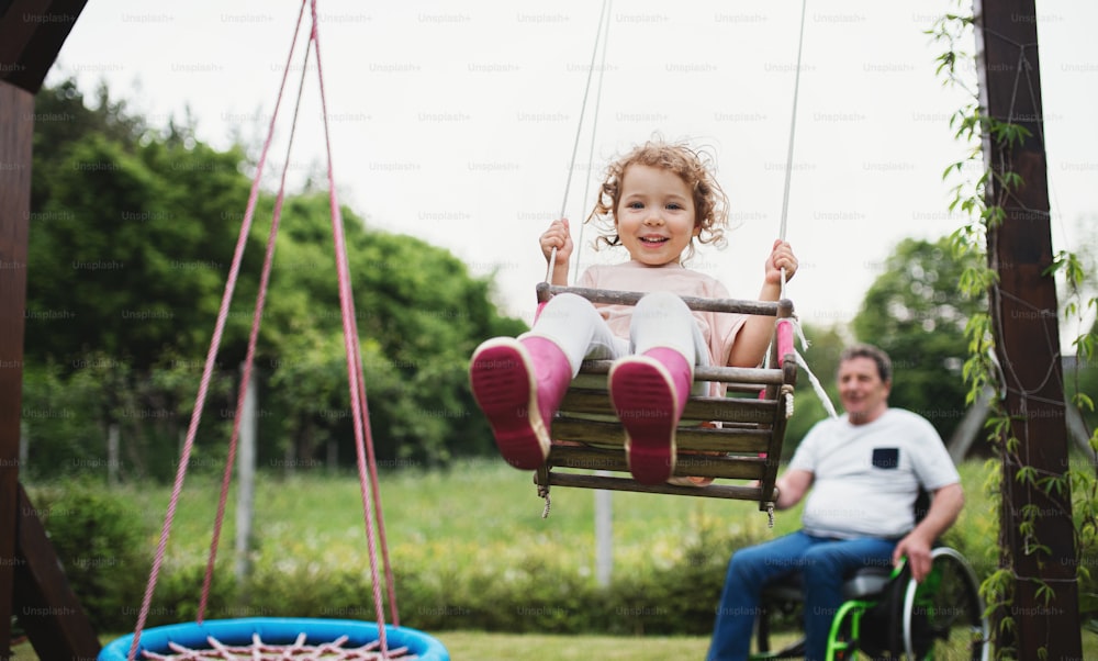 Piccola ragazza felice con il nonno anziano in sedia a rotelle che gioca nel giardino del cortile, oscillando.