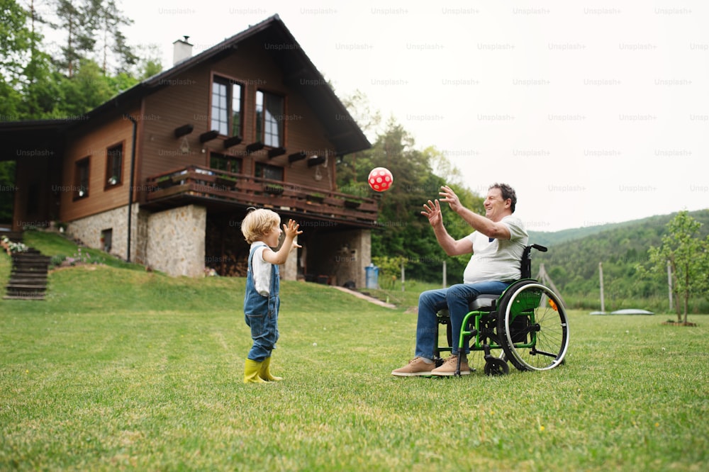 Niño pequeño feliz con abuelo mayor en silla de ruedas en el jardín, jugando con una pelota.