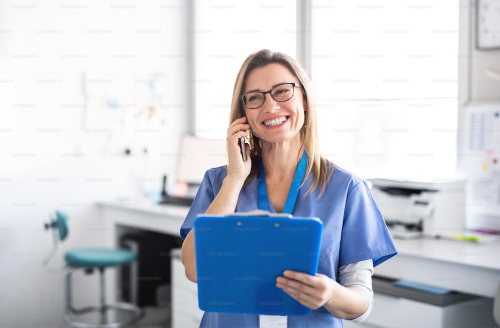 Un retrato de asistente dental alegre en la cirugía dental moderna, usando un teléfono inteligente.