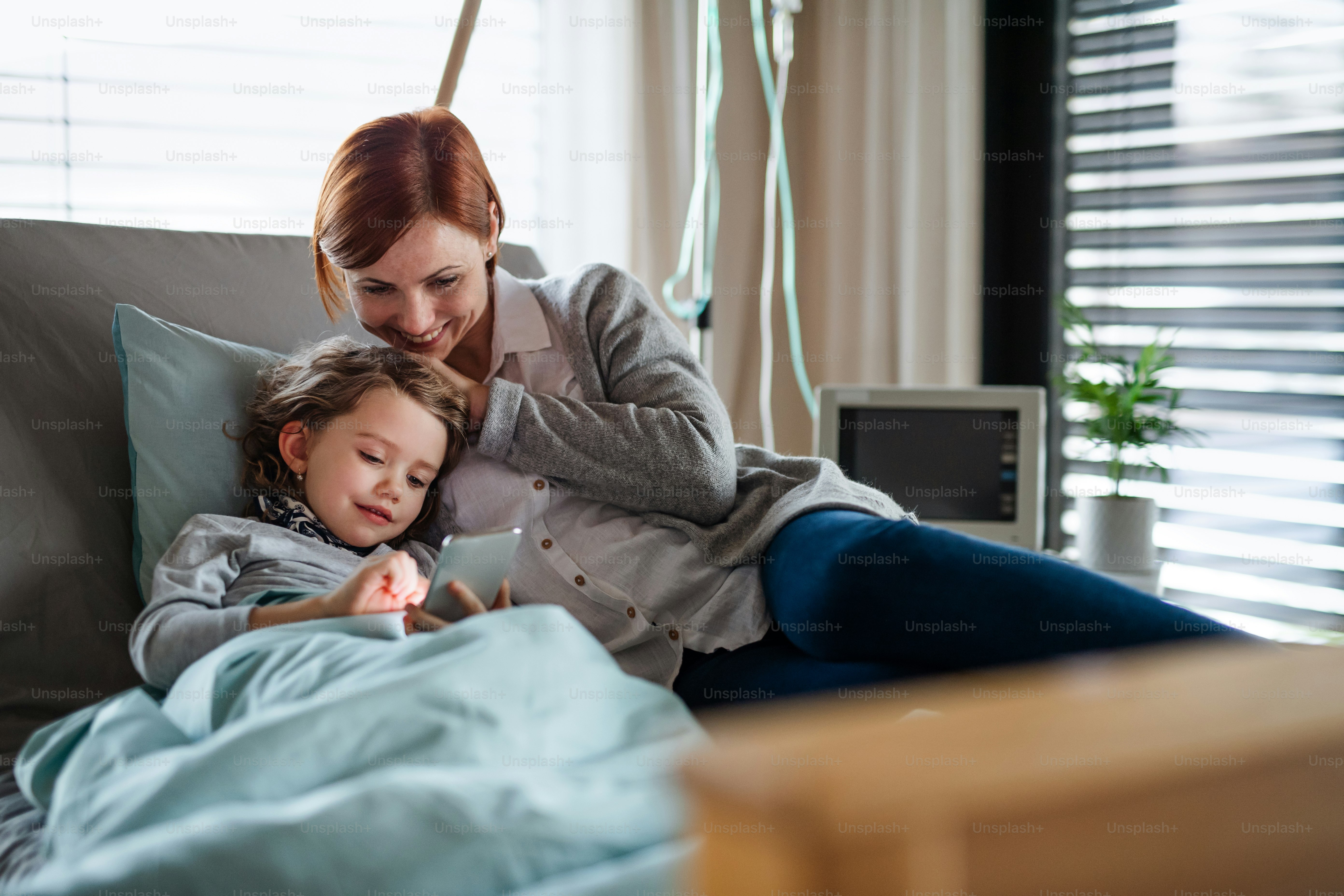 Happy small girl with mother in bed in hospital, using smartphone to pass time