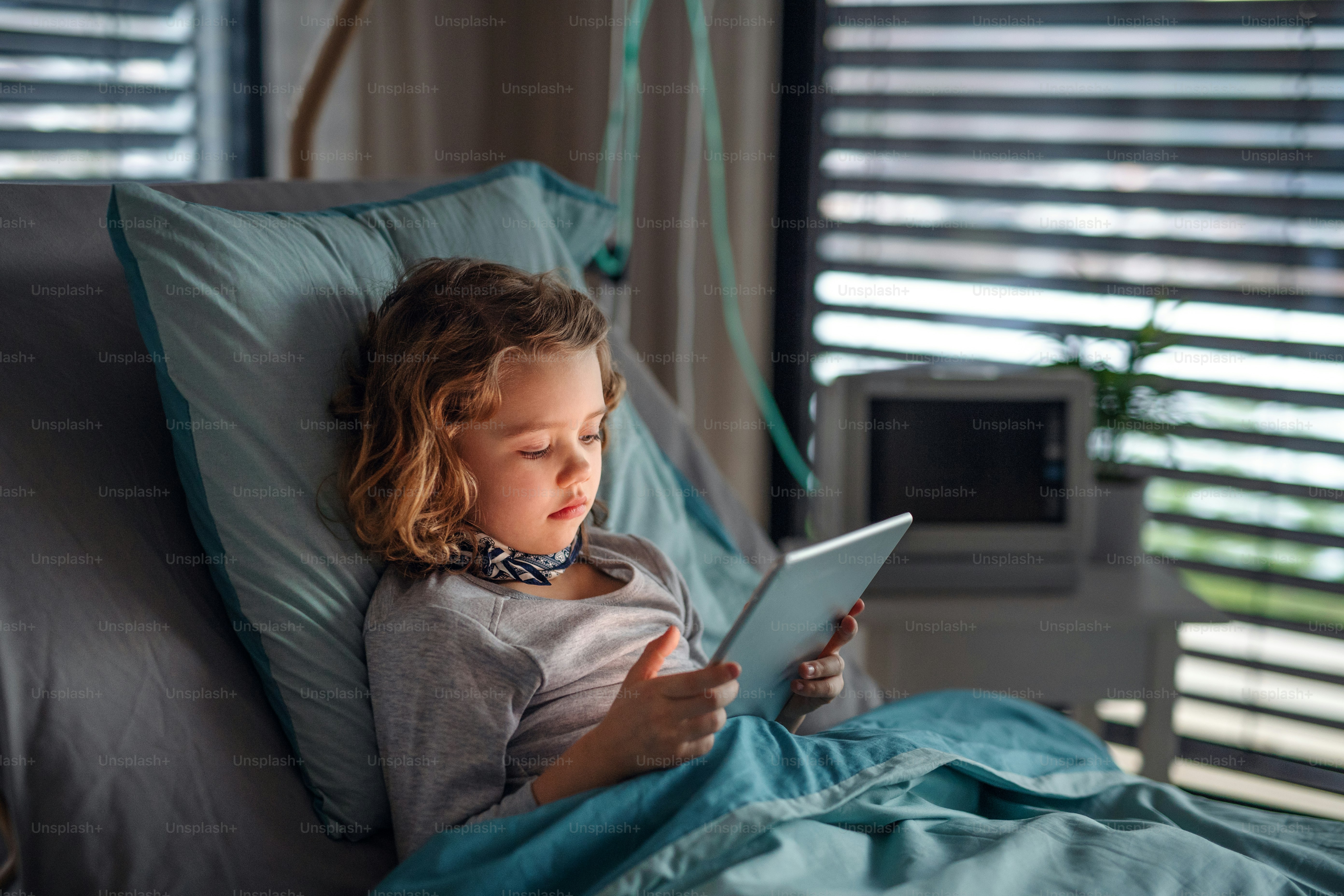 Bored small girl lying in bed in hospital, using tablet