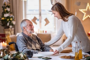 Jeune femme avec grand-père aîné en fauteuil roulant à l’intérieur à la maison à Noël, à la table.