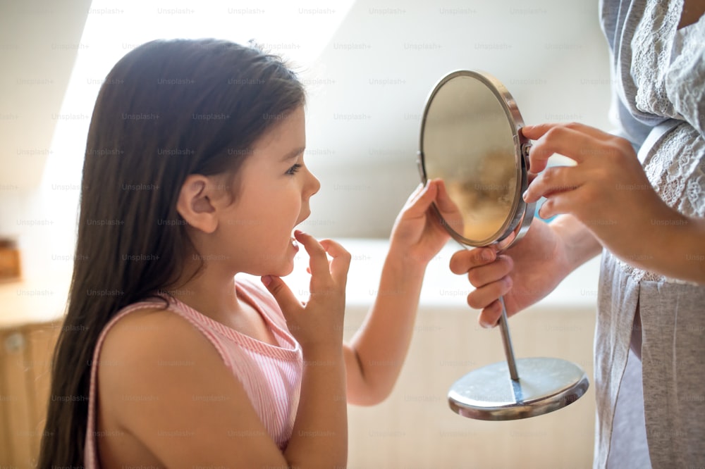 Retrato de niña pequeña con madre en el baño interior, mirando en el espejo al diente suelto del bebé.