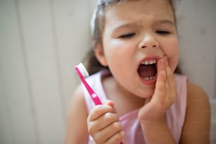 Front view portrait of worried small girl with toothbrush indoors, loosing baby tooth.