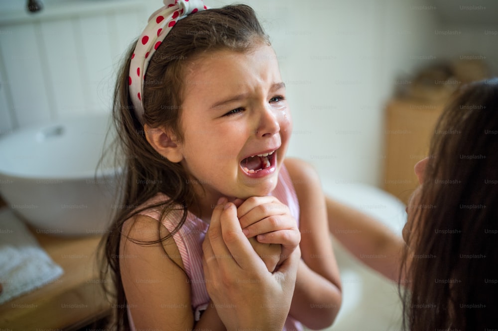 A cying small girl with unrecognizable mother indoors, loosing baby tooth.