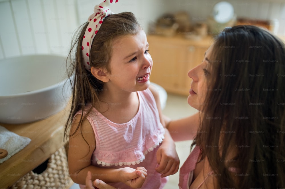 A cying small girl with mother indoors, loosing baby tooth.