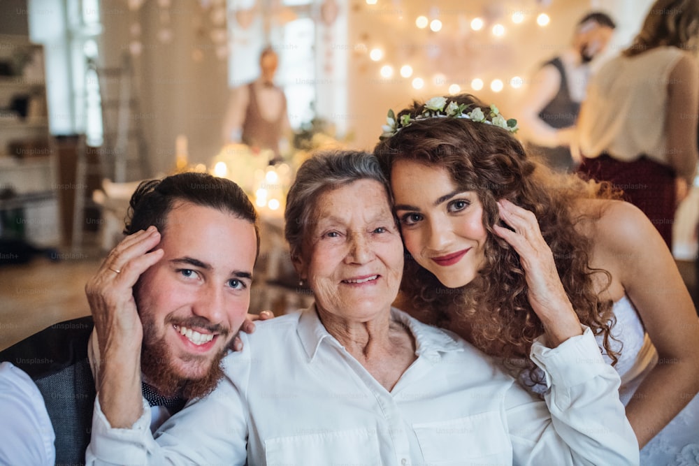 Ein junges Brautpaar mit Großmutter auf einer Hochzeitsfeier und schaut in die Kamera.