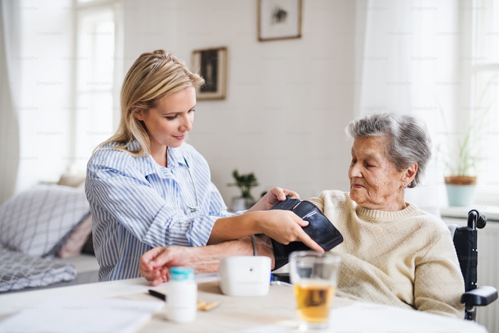 Un giovane visitatore sanitario che misura la pressione sanguigna di una donna anziana in sedia a rotelle a casa.