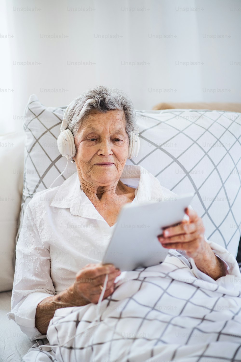 Eine kranke ältere Frau mit Kopfhörern und Tablet, die zu Hause oder im Krankenhaus im Bett liegt und Musik hört.