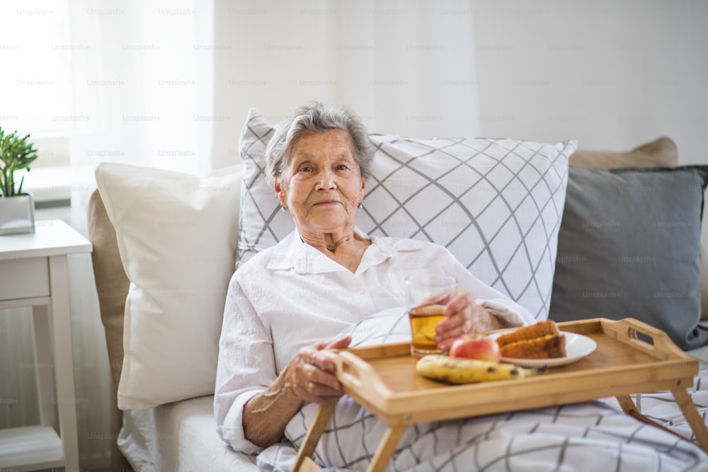 An elderly sick senior woman with food on a wooden tray lying in bed at home.