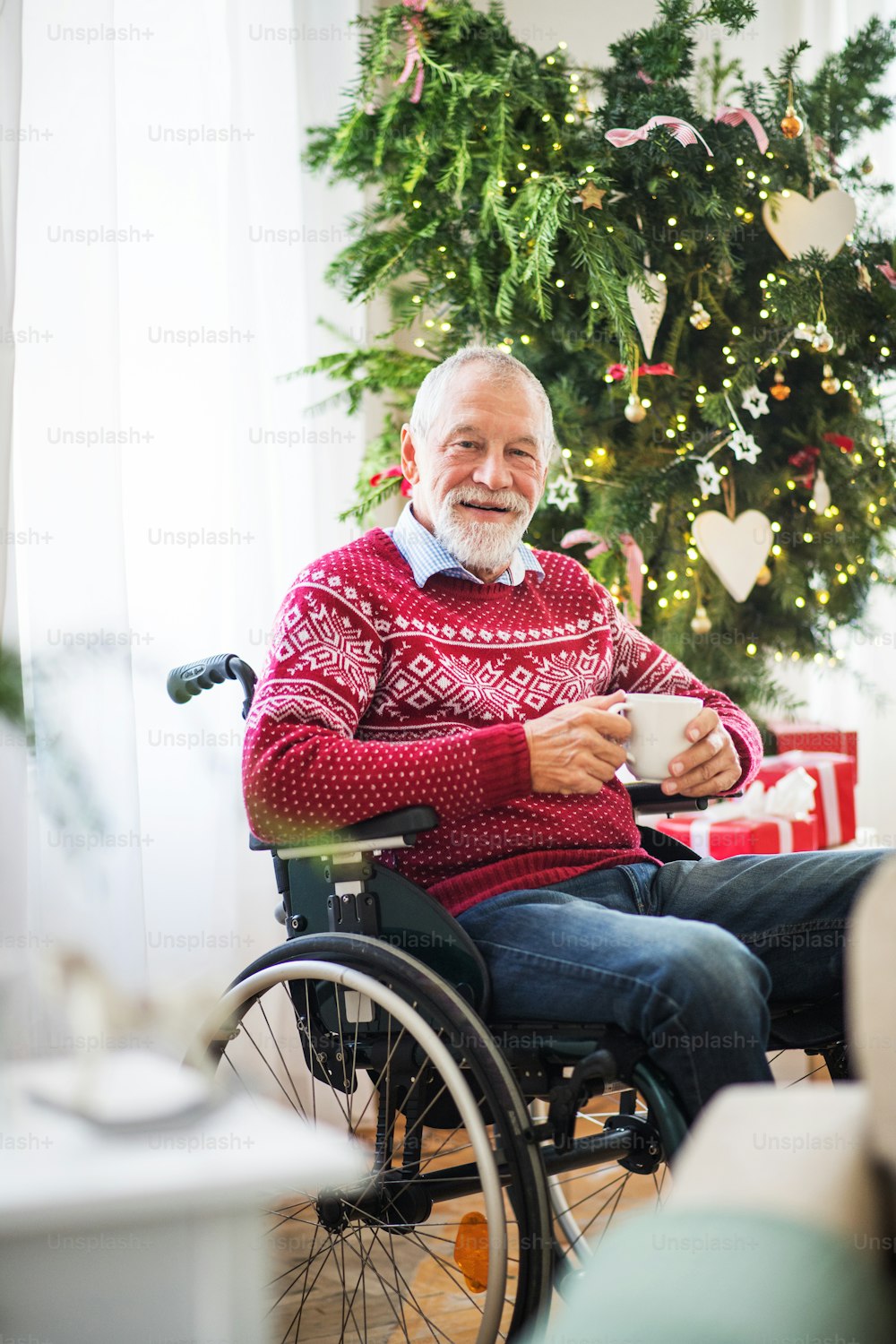 Un uomo anziano in sedia a rotelle con una tazza di tè o caffè a casa nel periodo natalizio.