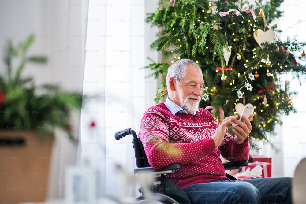 A senior man in wheelchair with a mobile phone at home at Christmas time, texting.