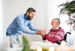 A hipster man giving a drink in a cup to his senior father in wheelchair at Christmas time.