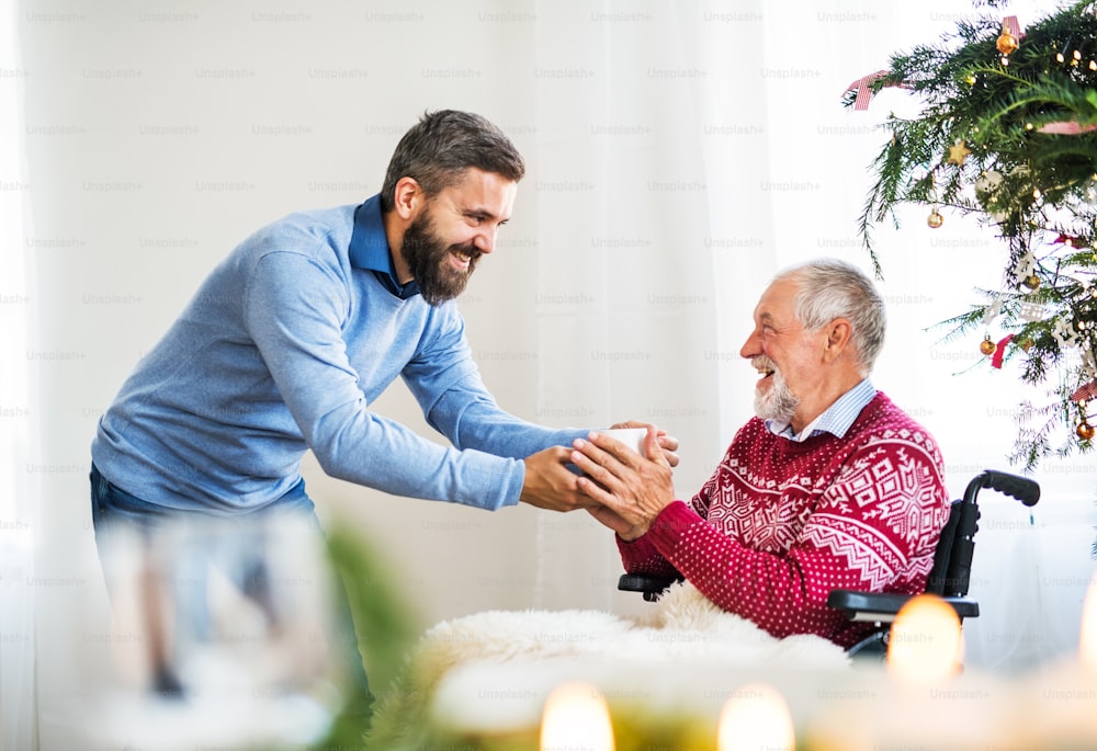 A hipster man giving a drink in a cup to his senior father in wheelchair at Christmas time.