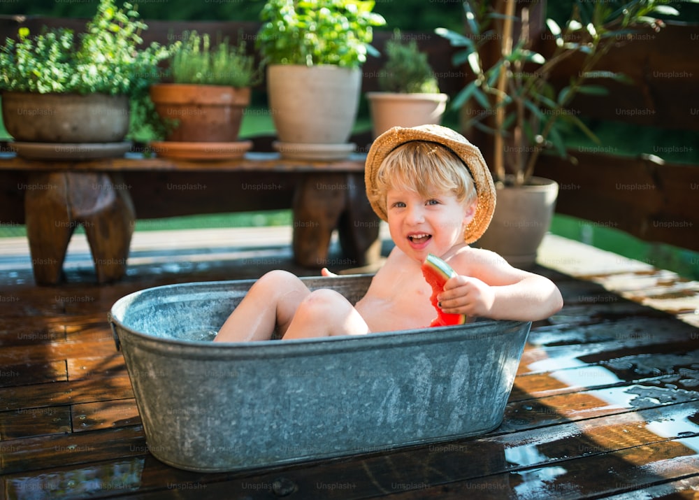 Happy small boy with a hat in bath tub outdoors in garden in summer, eating watermelon.